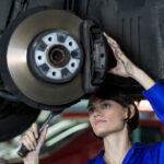 A person in a blue work uniform is using a wrench to repair a vehicle's brake assembly, seen from below the car.