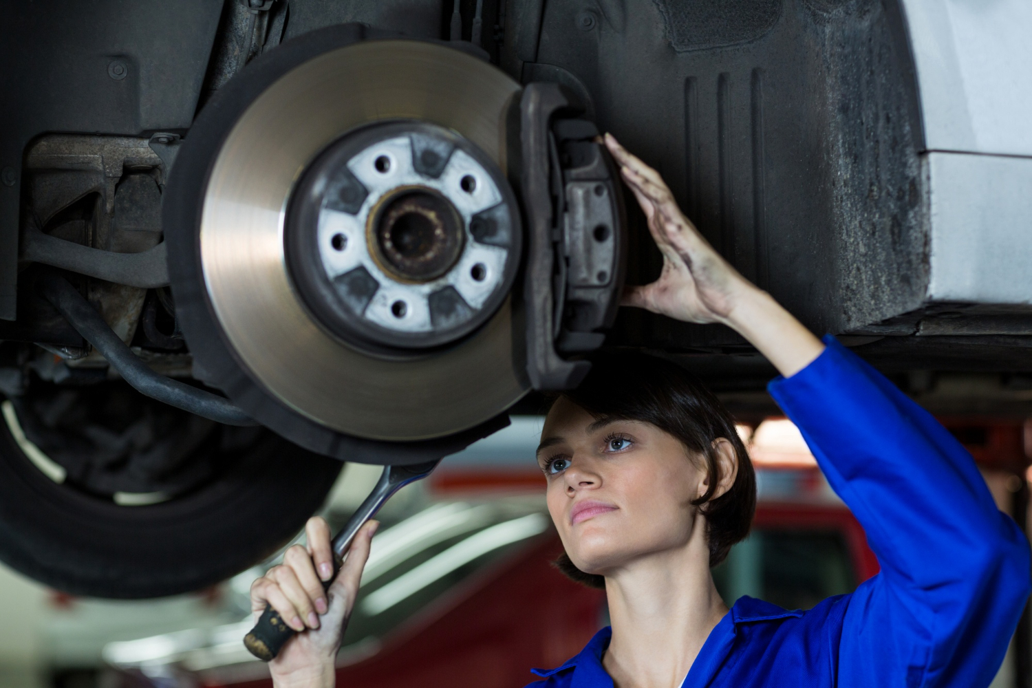 A person in a blue work uniform is using a wrench to repair a vehicle's brake assembly, seen from below the car.