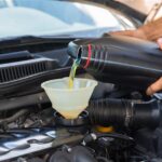 A person pours motor oil from a black bottle into a funnel placed in an engine bay, demonstrating vehicle maintenance.