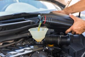A person pours motor oil from a black bottle into a funnel placed in an engine bay, demonstrating vehicle maintenance.