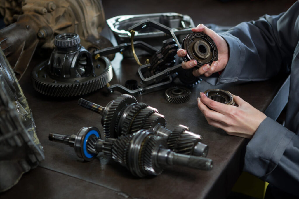 A person in a gray work shirt examines mechanical gears on a workbench, surrounded by various disassembled automotive components.