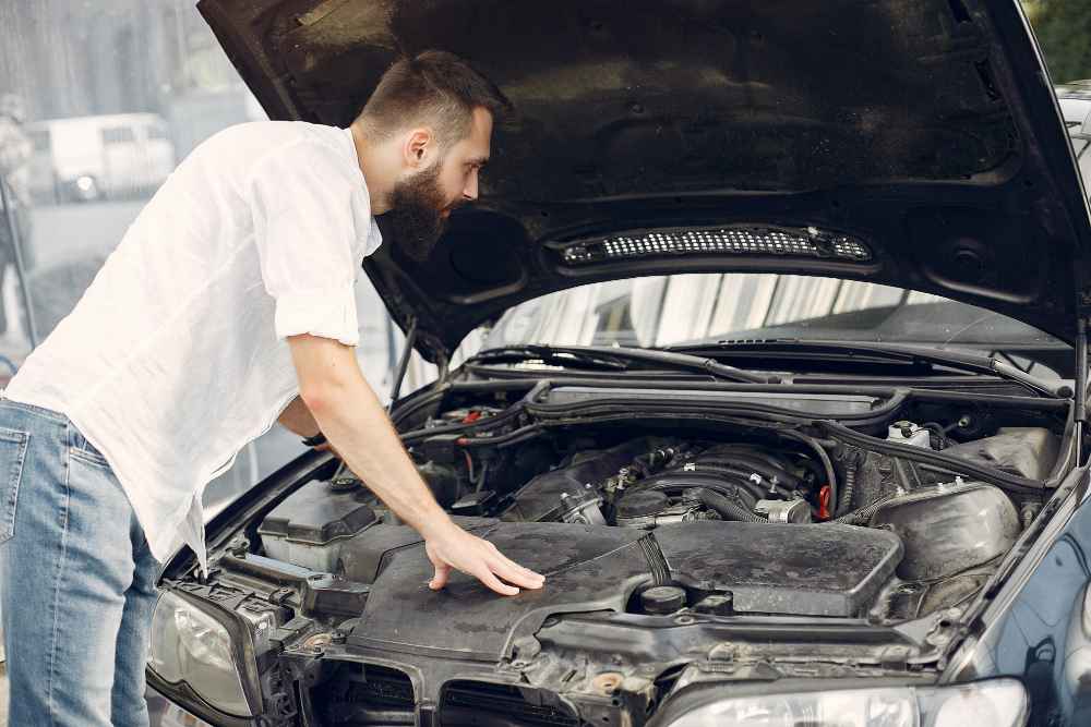 A person inspects the engine of a car with the hood raised, dressed in a white shirt and blue jeans.