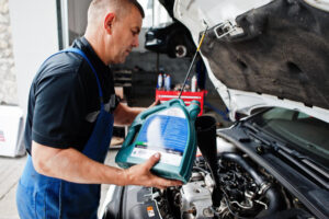 A mechanic in blue overalls pours engine oil into a funnel above an open car hood, with tools and equipment in the background.