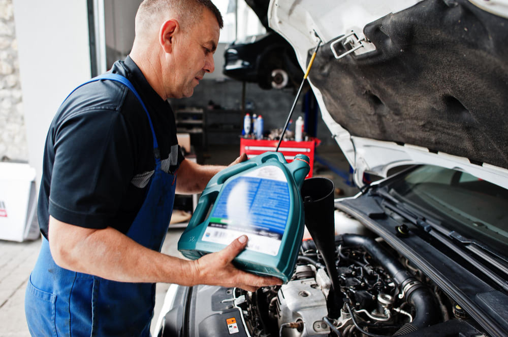 A mechanic in blue overalls pours engine oil into a funnel above an open car hood, with tools and equipment in the background.
